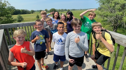 around 11 children smiling and giving the thumbs up on a boardwalk with nature background