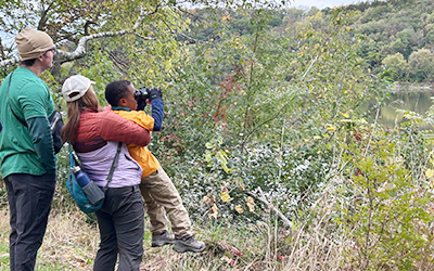 A family looking standing at an overlook. One parent is holding up a child and the child has binoculars and is looking through them.